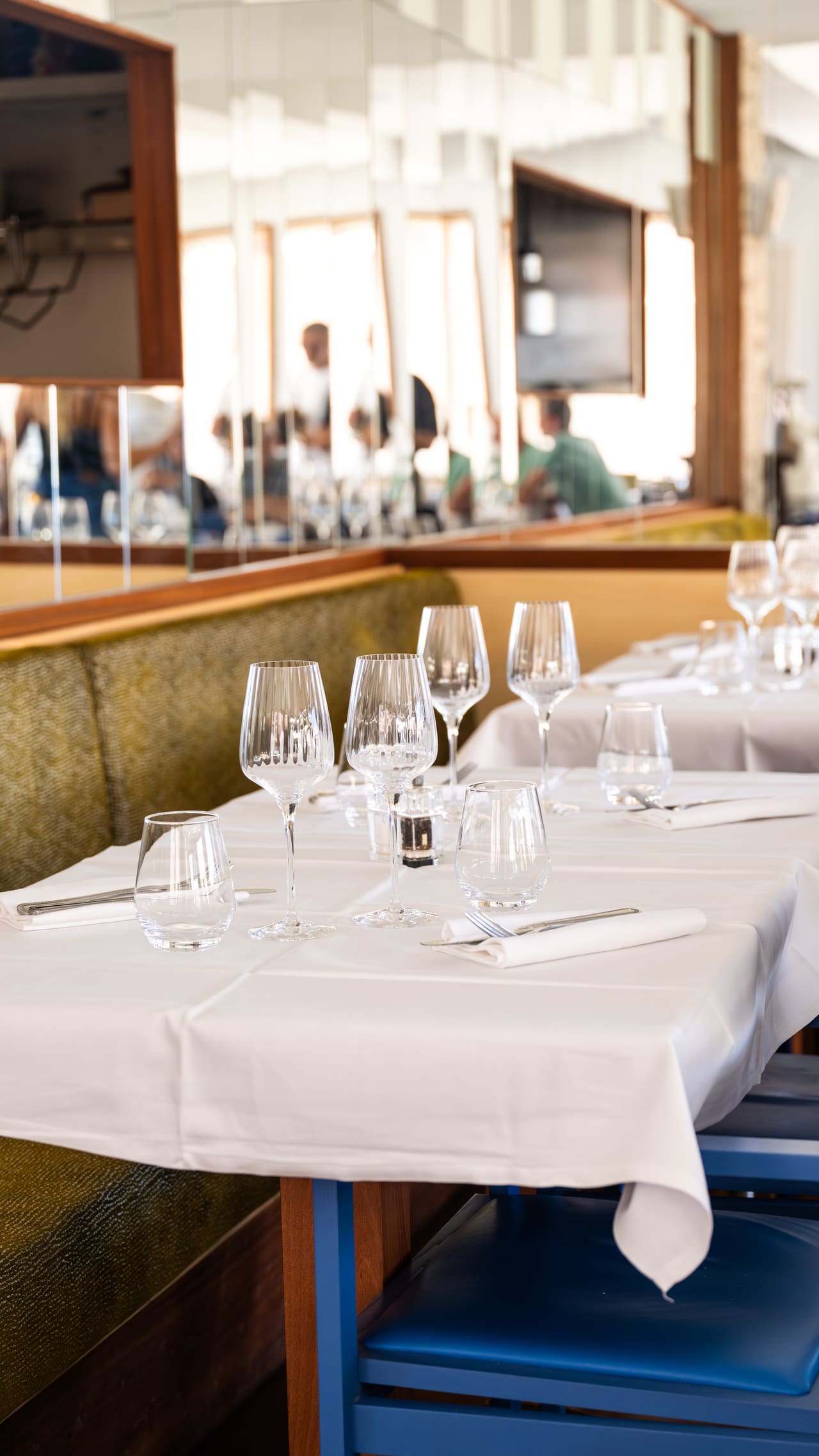 Une table dressée en salle : nappe blanche, verres au soleil, banquette et chaise bleue